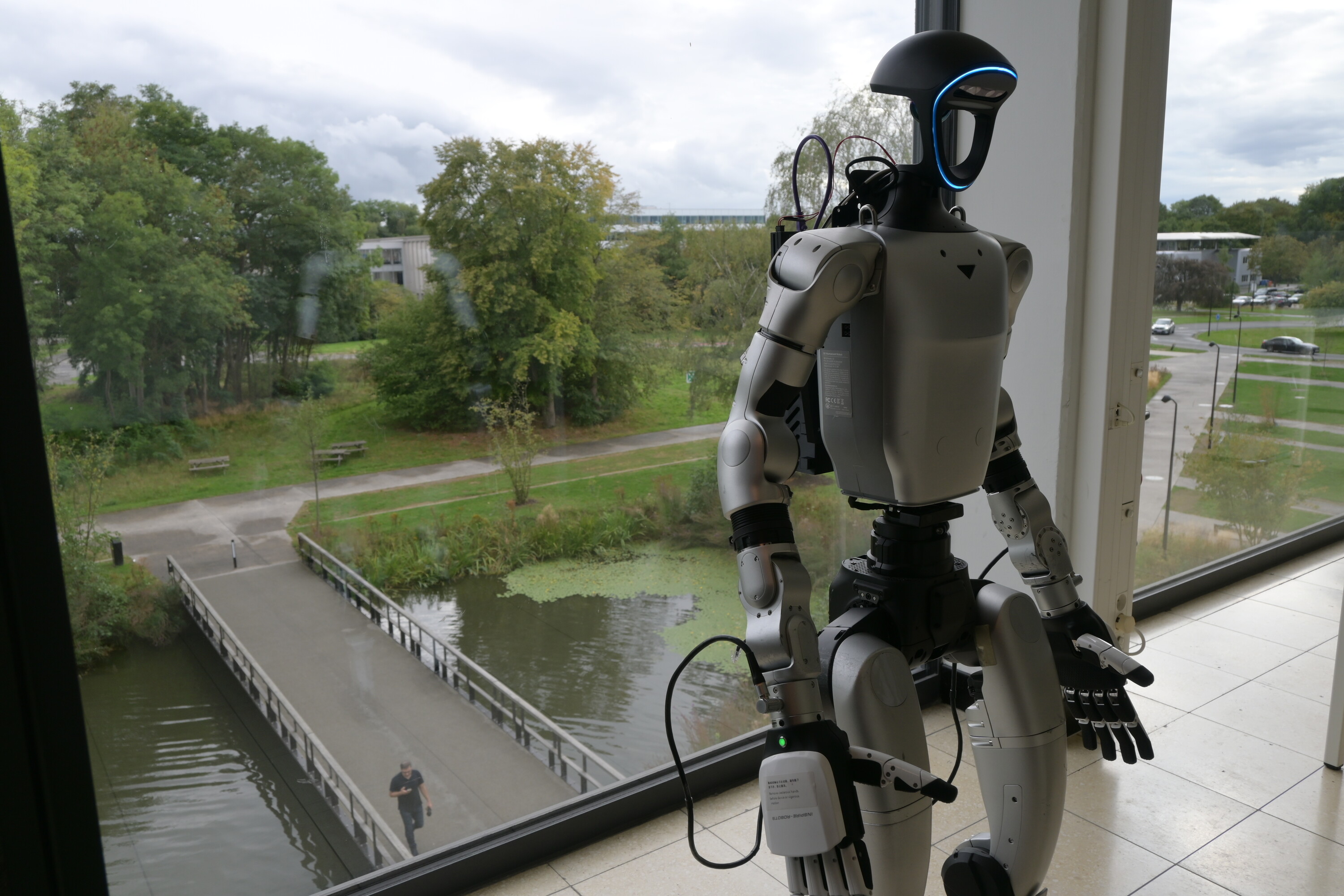 Unitree G1 humanoid standing by a window overlooking a canal and trees.